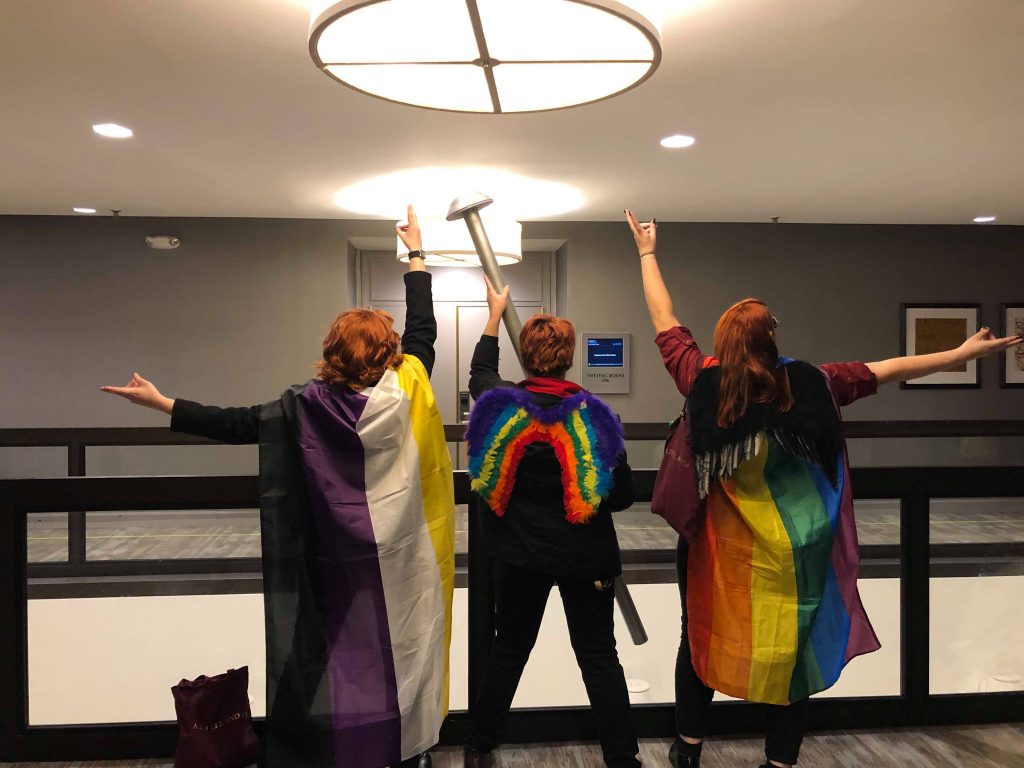 Three androgynous Crowley cosplayers look over a balcony at Dragon Con, dramatically posing for a coming out party portrait. Two are wearing pride flags as capes. The middle one has rainbow wings and a six-foot tall pin prop.