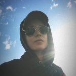 An androgynous person wears sunglasses, a baseball cap, and a hoodie with the hood up. Behind them is a blue sky with small clouds and bright sunlight