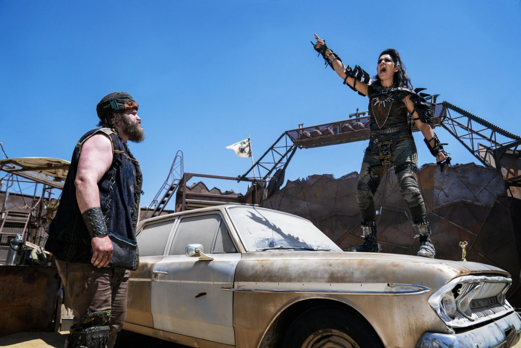 a female road warrior with fist upraised stands on a burned out car