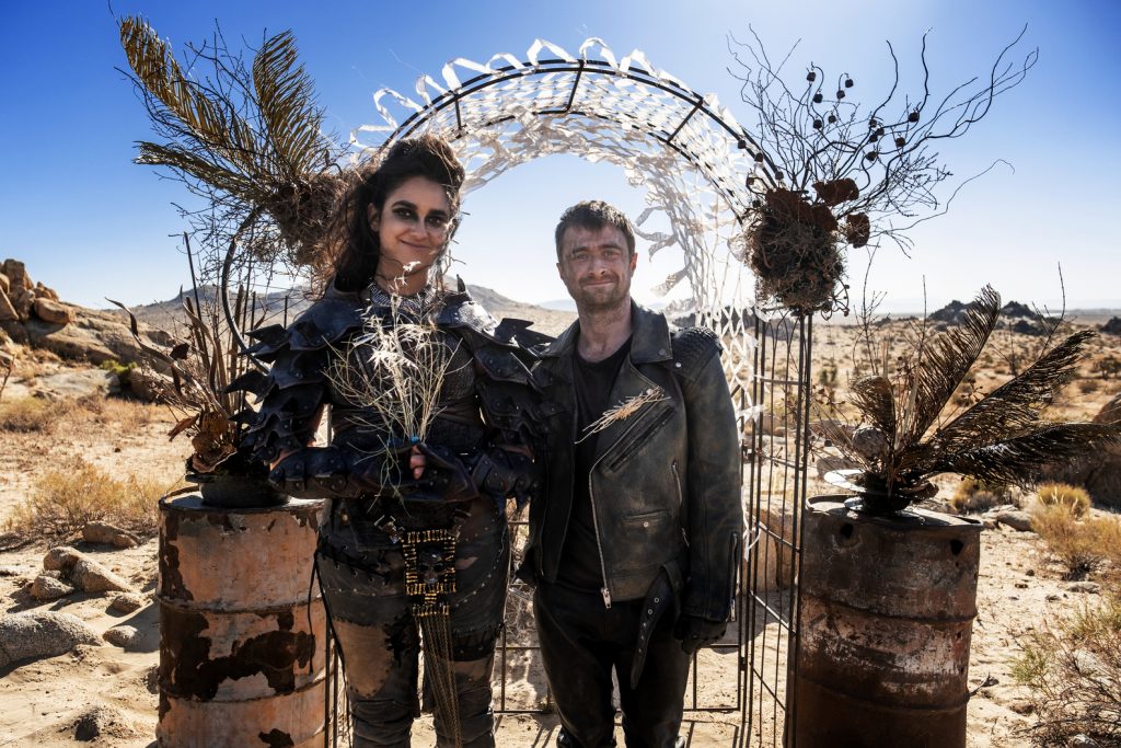 a woman dressed in road-warrior style clothing standing next to a man in apocalypse-leather, in front of a wedding arch
