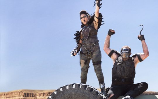 a woman in road warrior outfit raises her hand in celebration