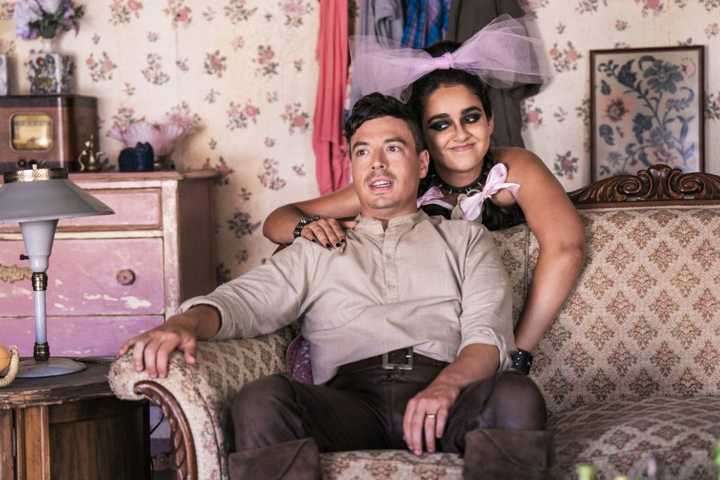 a man sitting on a couch and a woman with a pink bowtie standing behind him