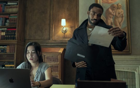 Donald Glover stands, looking at a marriage certificate, while Maya Erskine sits at the desk looking up something on a computer