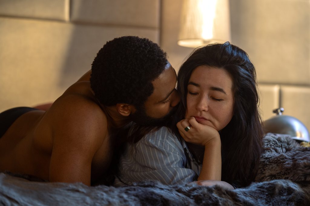 Donald Glover and Maya Erskine in bed, Donald snuggling with Maya.