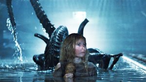 A little girl with messy hair, Newt, in water with an Alien coming up behind her, tail in the air.