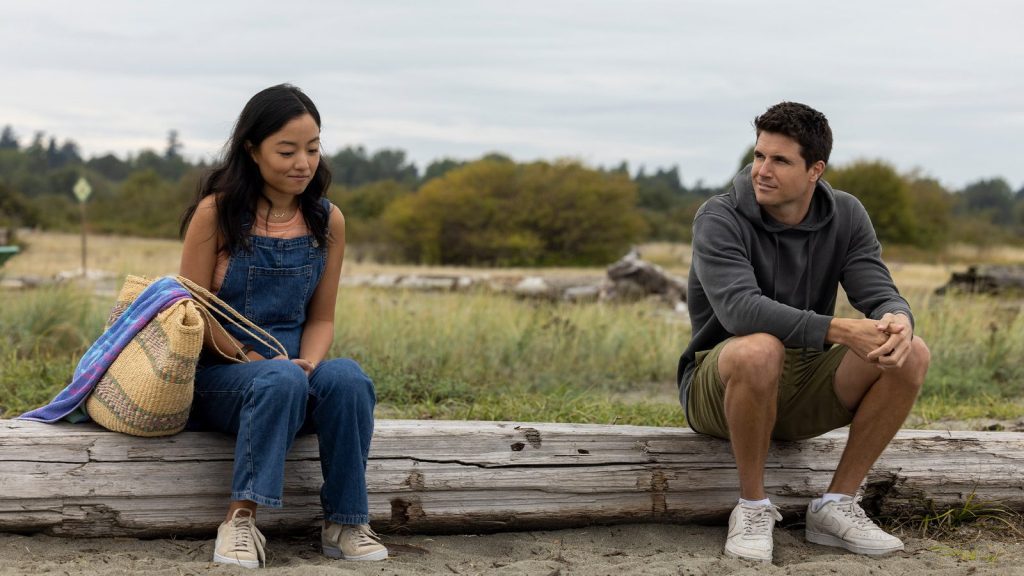 Andrea Bang in denim overalls and Robbie Amell in sweatpants and shirt sit on a wooden wall.