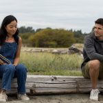 Andrea Bang in denim overalls and Robbie Amell in sweatpants and shirt sit on a wooden wall.