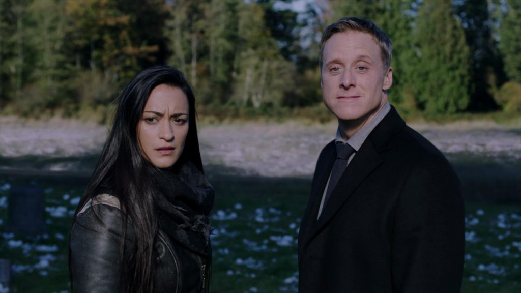 a Native American woman in a brown jacket on the left, and a white male in a black suit look at the camera in front of a river