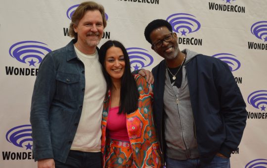 a tall white man in a denim shirt over a white shirt; a young Native American woman in a multi-colored outfit; and a black man in a blue jacket over a gray shirt