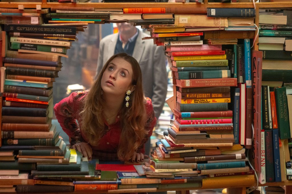A Wall of books stacked sideways with a hole in the center, out of which a young white woman peers through 