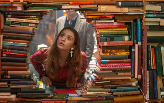 A Wall of books stacked sideways with a hole in the center, out of which a young white woman peers through
