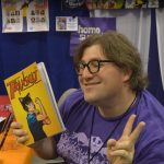 a young trans woman with brown hair and glasses, wearing a purple shirt. One hand has a TransCat comic in it and the other hand is making the peace sign.