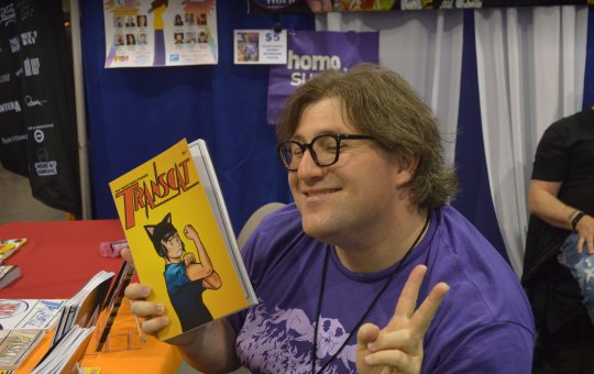 a young trans woman with brown hair and glasses, wearing a purple shirt. One hand has a TransCat comic in it and the other hand is making the peace sign.