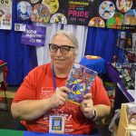 An older woman with white hair and glasses, wearing an orange shirt, holds a copy of a graphic novel in a booth at WonderCon