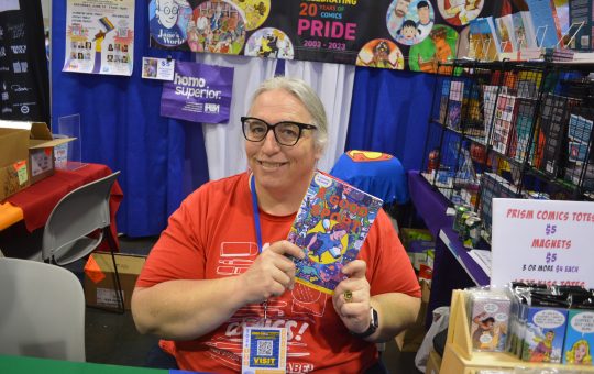 An older woman with white hair and glasses, wearing an orange shirt, holds a copy of a graphic novel in a booth at WonderCon