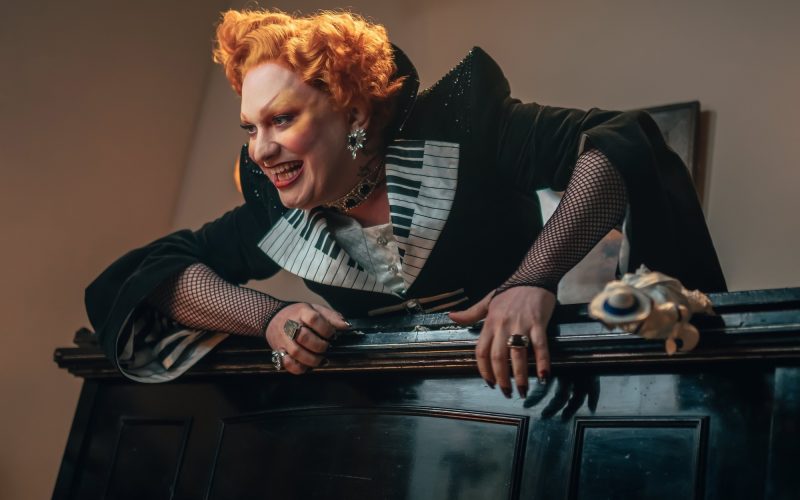 a person with red hair and a biano-themed top crawls out of a piano
