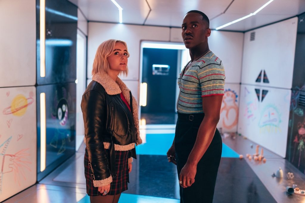 a young white woman in pleather jacket and a youngish black man in a striped shirt stare just above the camera in a corridor