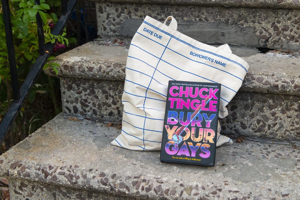 A copy of Chuck Tingle's 2024 release Bury Your Gays sits on a step. Behind it is a bag with a library card design. 