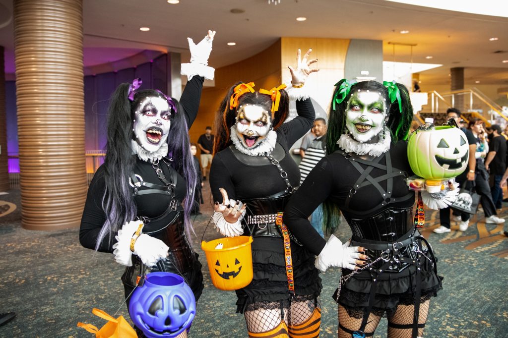 three women, dressed in black outfits and wearing white makeup, pose with trick or treat containers