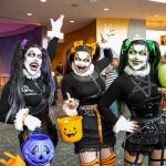 three women, dressed in black outfits and wearing white makeup, pose with trick or treat containers