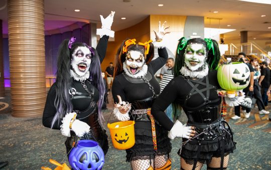 three women, dressed in black outfits and wearing white makeup, pose with trick or treat containers