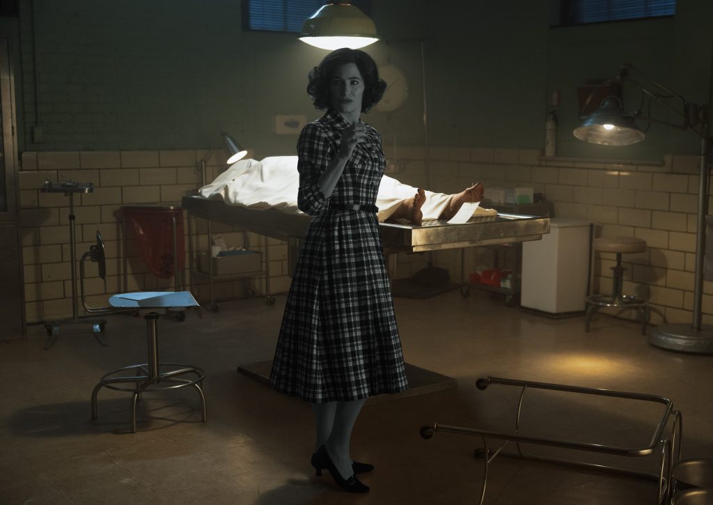 an autopsy room with a body under a sheet on a bed/table. Agatha (Katherine Hahn) is in black and white and 60's style clothes, looking at someone.