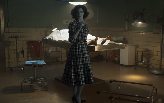 an autopsy room with a body under a sheet on a bed/table. Agatha (Katherine Hahn) is in black and white and 60's style clothes, looking at someone.