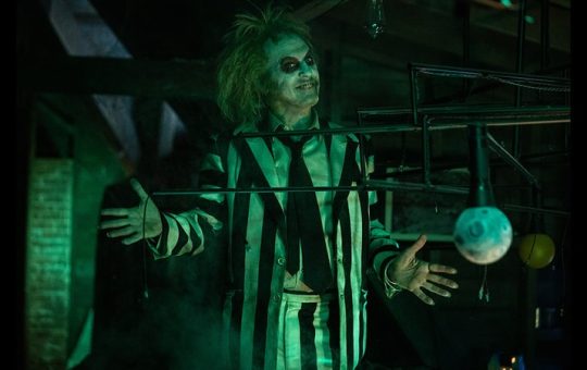 Michael Keaton as Betelgeuse - white and black striped suit, clown white makeup, and wild hair - posing with both hands out