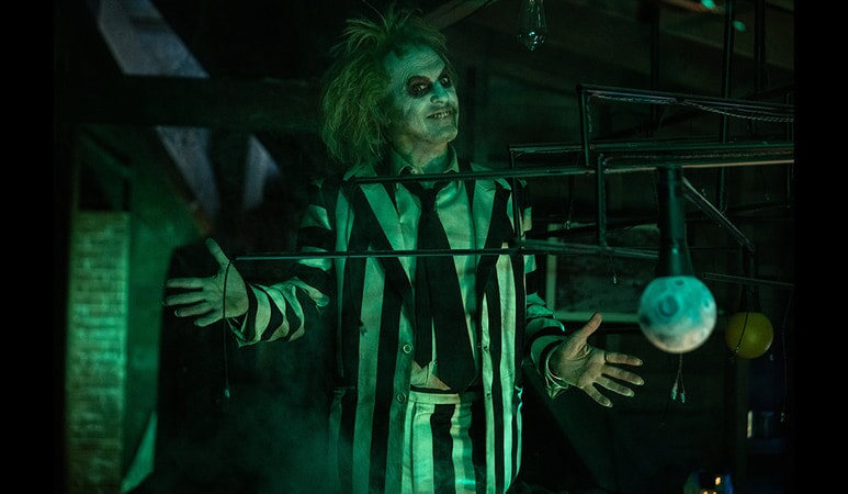 Michael Keaton as Betelgeuse - white and black striped suit, clown white makeup, and wild hair - posing with both hands out