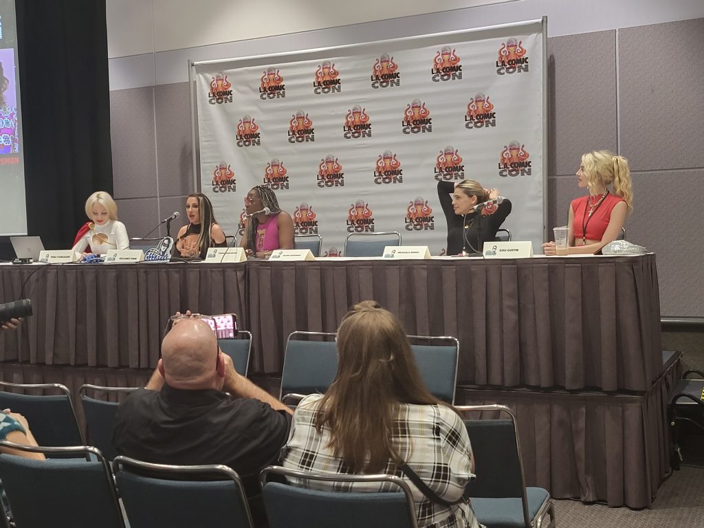 Five women sit at a table during a panel. The LA Comic Con banner is behind them. 