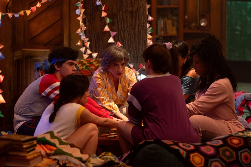 Joe Locke, Patti LuPone, Alice Wu-Gulliver, Sasheer Zamata, Hathryn Hahn, and Aubrey Plaza sitting in a circle in 80s clothing around a Ouija board. 