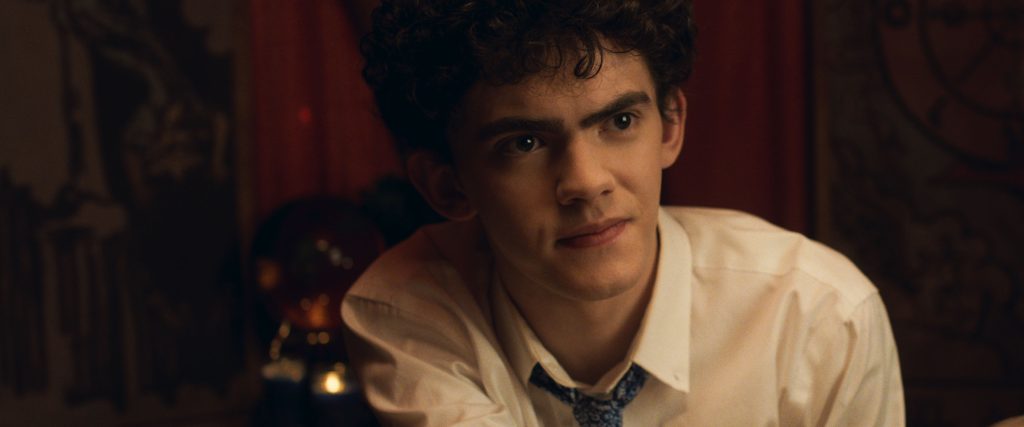 A white teen boy (Joe Locke) with black curly hair and a button up shirt and loose tie, sits looking curious. 