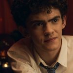 A white teen boy (Joe Locke) with black curly hair and a button up shirt and loose tie, sits looking curious.