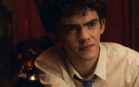 A white teen boy (Joe Locke) with black curly hair and a button up shirt and loose tie, sits looking curious.