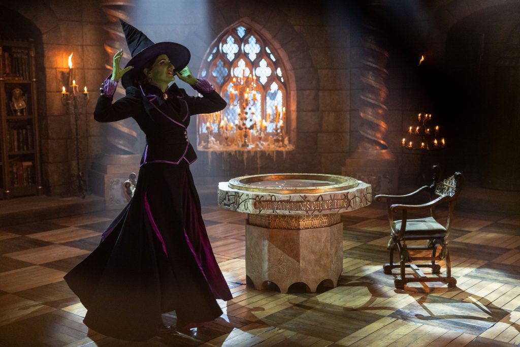 A woman wearing green make up and a black witch outfit (Kathryn Hahn) stands next to a table in a castle. 