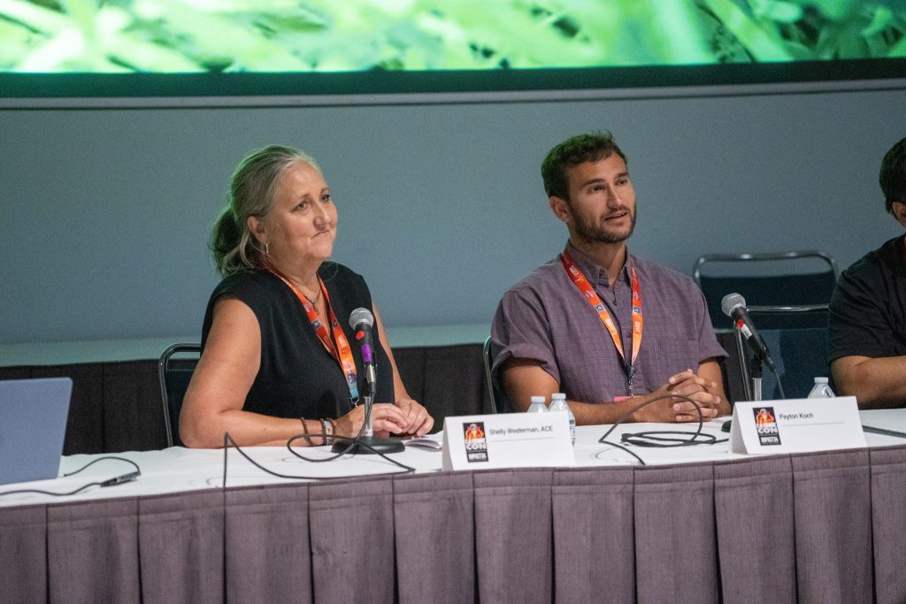 An older woman with blond-white hair and a black top on the left and a younger man with black hair and wearing purple shirt sit at a table for a panel.