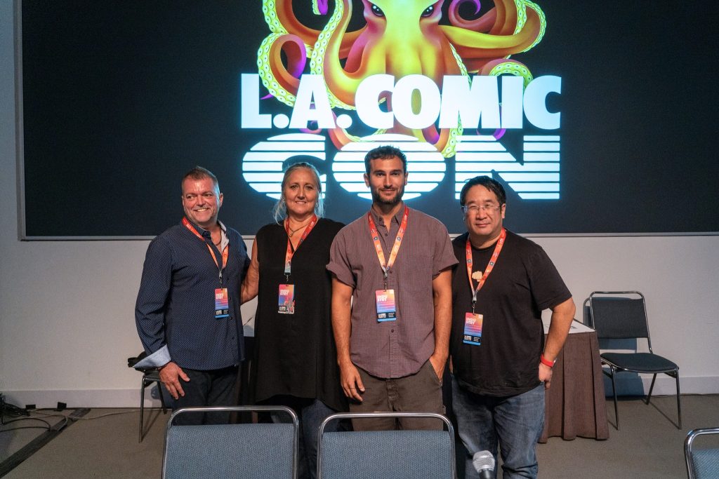 Four people stand in a room with the LA Comic Con logo behind them. From left to right: a middle-aged white man with a crew cut in a dark blue shirt, an older woman with white-blond hair and a black shirt, a younger man in a light purple shirt, and another middle-aged man in a black shirt. 