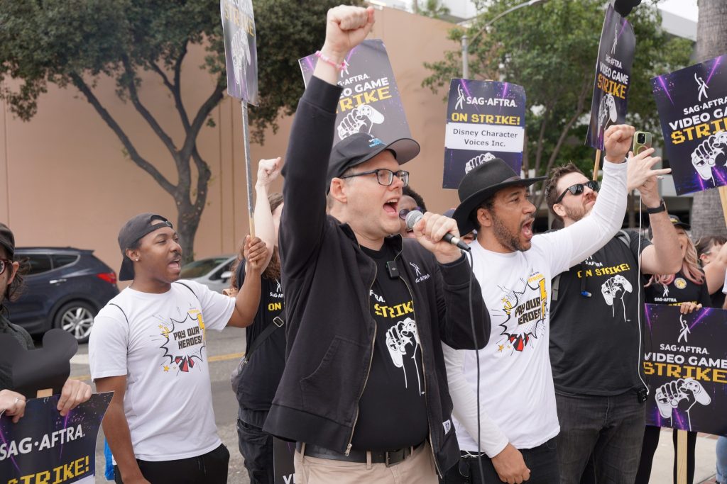 a crowd of people at a SAG-AFTRA strike line, one person with a microphone and their hand in the air