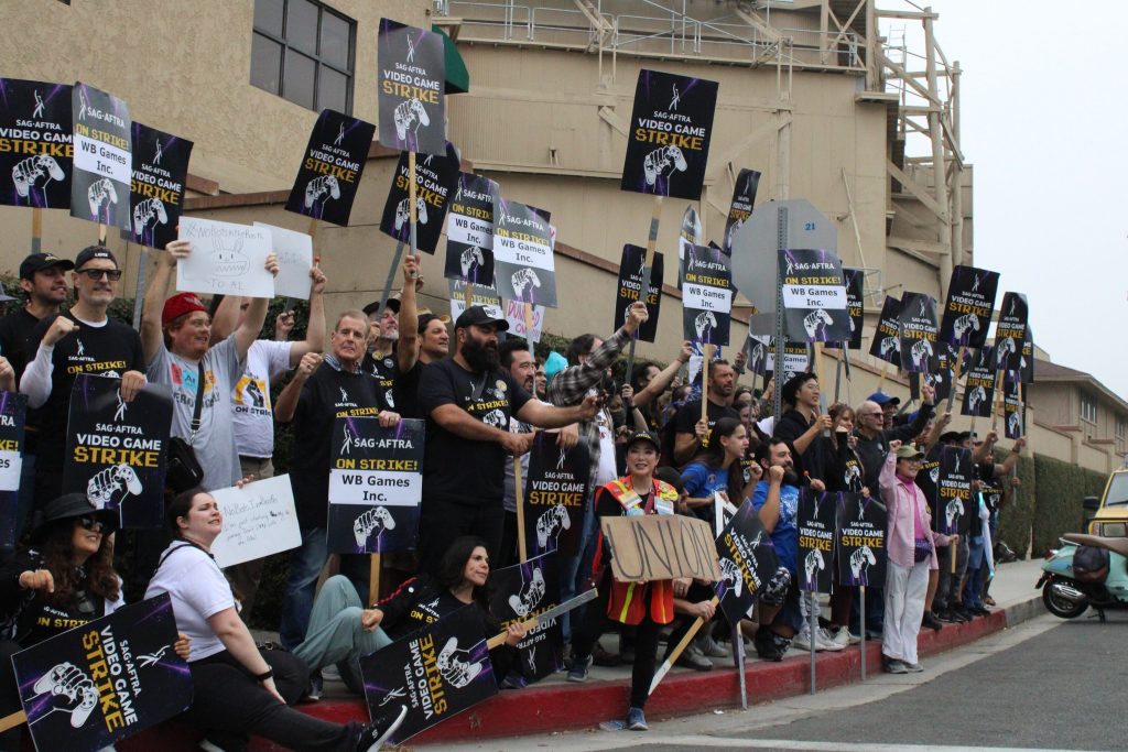 a crowd of people on a sidewalk with signs for the SAG-AFTRA video game strike