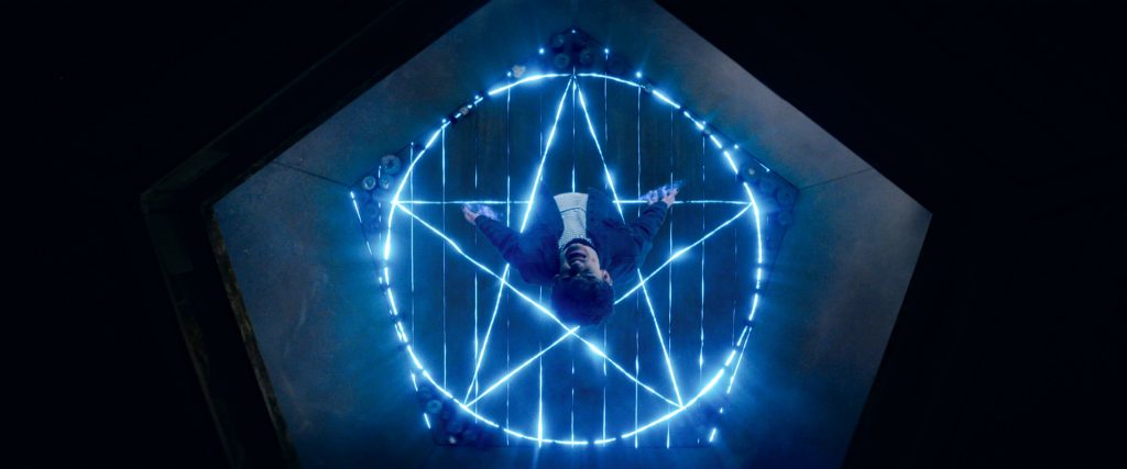 an overhead shot of a teenager with black hair and a blue and white outfit standing on a floor with an electric blue pentagram.