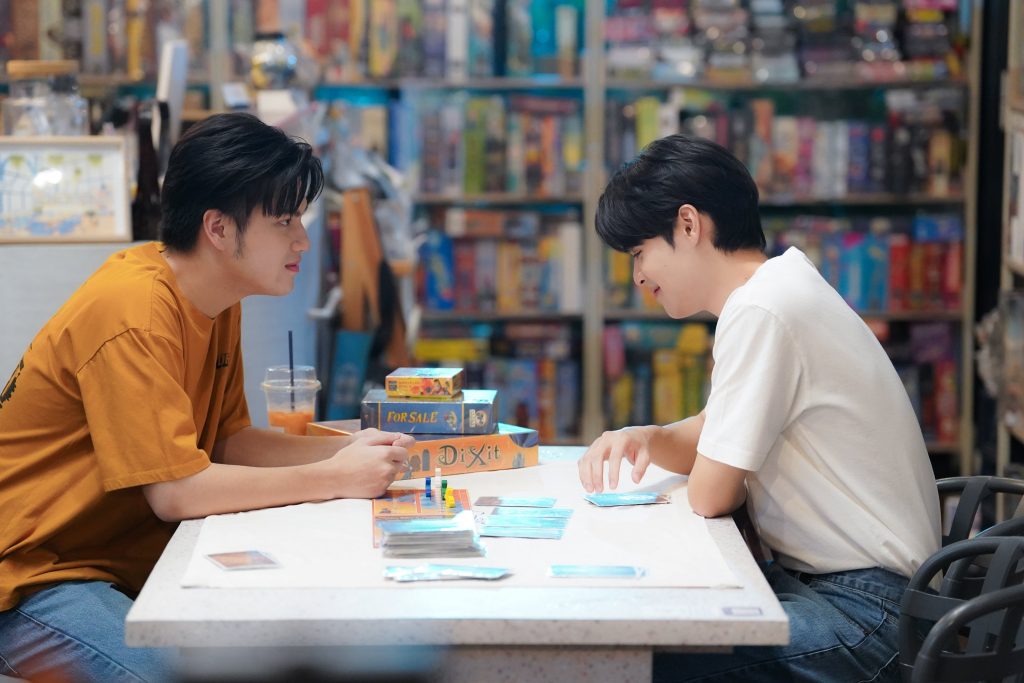 Pond and Sand sit across the table from each other at a board game cafe. An abandoned game is on the table. Sand is smiling down at the table. Pond is smiling at Sad.