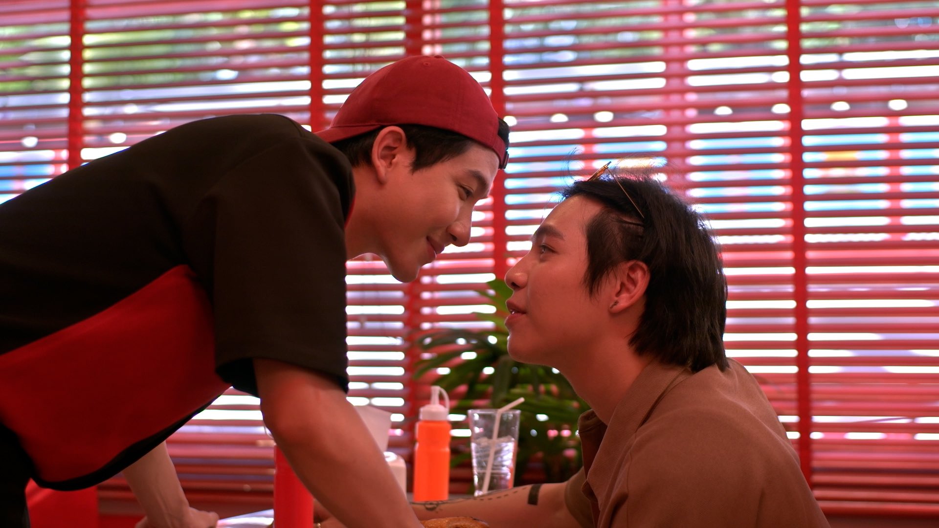 Kant and Bison are in Heart Burger, an American-style diner. Kant is seated in a booth, leaning on the table and smiling up at Bison. Bison is in his uniform (with backwards cap), both hands braced on the table, leaning down towards Kant and smiling.