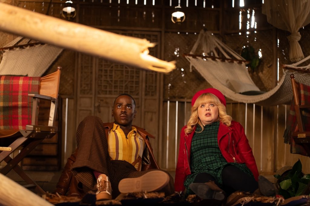 a black man in a brown jacket, striped shirt, and brown pants lies on his elbows on the floor next to a white woman with blond hair wearing a red hat and jacket and a green plaid dress. They are in a 'adventure' type cabin.