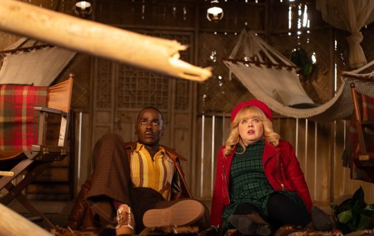 a black man in a brown jacket, striped shirt, and brown pants lies on his elbows on the floor next to a white woman with blond hair wearing a red hat and jacket and a green plaid dress. They are in a 'adventure' type cabin.