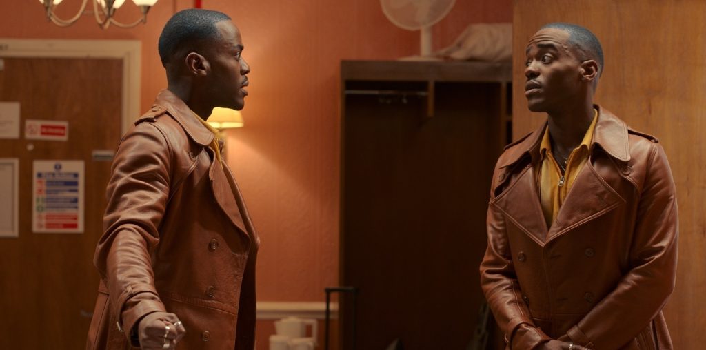 Two versions of the same black man, wearing a brown jacket, striped shirt, and brown pants, standing across from each other in conversation in a hallway.