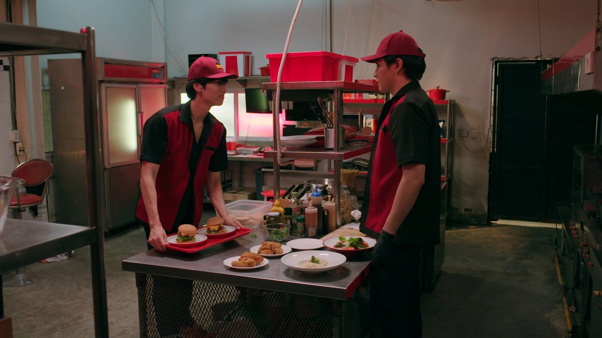 Style and Fadel stand across from each other in the Heart Burger kitchen. They are both wearing the uniform of black pants, black and red shirt, red cap. The table between them is filled with condiments, ingredients, and finished burgers. Style is picking up a tray with two burgers on it and smirking at Fadel.