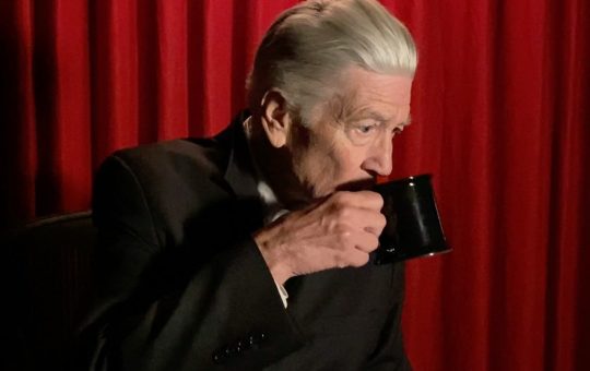 Filmmaker David Lynch drinks from a coffee cup in front of a red curtain.