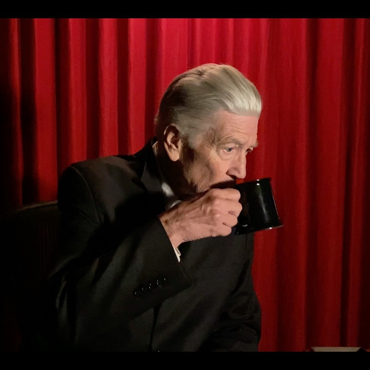 Filmmaker David Lynch drinks from a coffee cup in front of a red curtain.