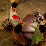 An overhead shot of a table in an abandoned building. Bison, Fadel, Kant, and Style are gathered around the table, standing up, with their hands overtop of each other's.