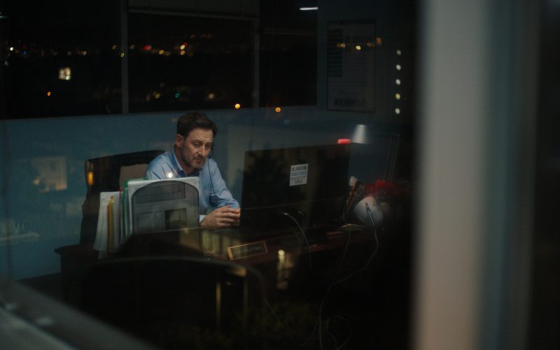 a reflection through a window: a white man with dark hair and a blue long-sleeved shirt sits at a desk.